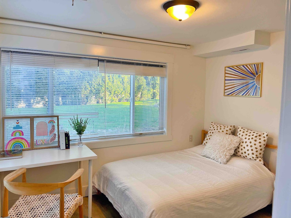 A bright bedroom features a spacious bed with a white coverlet. A small desk and chair are positioned nearby, accompanied by colorful framed artwork. Natural light fills the room through a large window overlooking greenery.