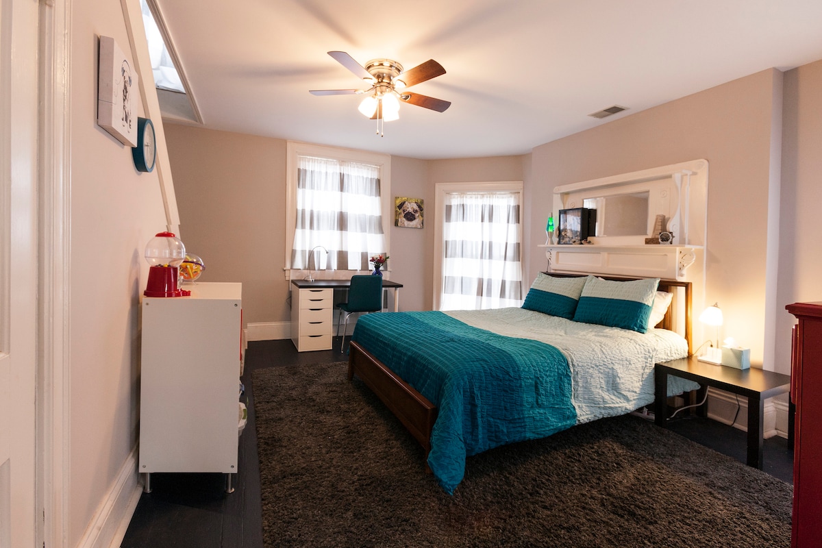 A spacious bedroom is furnished with a large bed covered in a soft turquoise quilt. Dual windows provide ample natural light, and a ceiling fan adds comfort. Nearby, a dresser and desk are positioned against the wall, all set upon a plush dark rug.