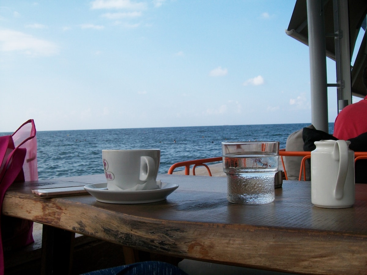 A table is set along the shoreline, featuring a small cup and saucer, a glass of water, and a milk jug. The expansive sea stretches out in the background under a light blue sky with a few clouds. The beachfront ambiance is calm and inviting.