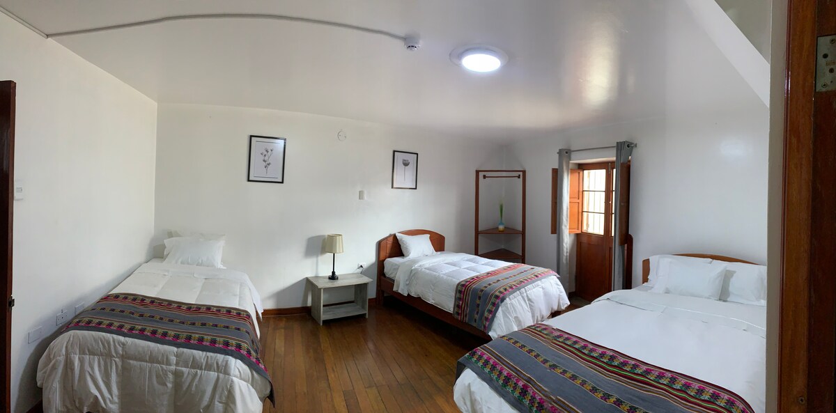 A spacious room is furnished with three beds featuring white linens and colorful woven accents. A small bedside table with a lamp stands between two beds. Natural light enters through large windows dressed with sheer curtains, illuminating the room's wooden flooring and minimalist decor.