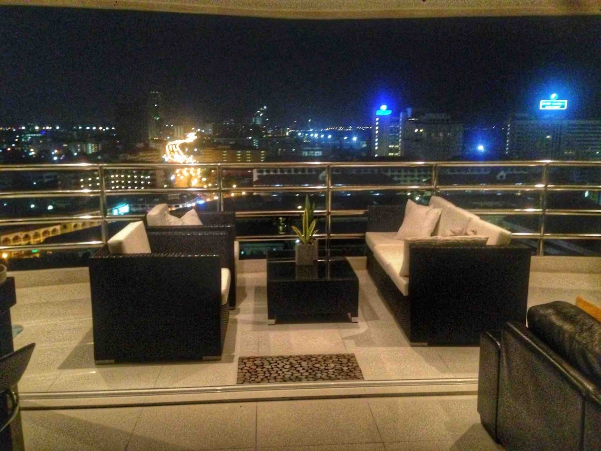 A stylish balcony is showcased with modern seating, including two armchairs and a sofa surrounding a small table. City lights illuminate the night view, offering a glimpse of urban activity. A decorative plant adds a touch of greenery to the space.