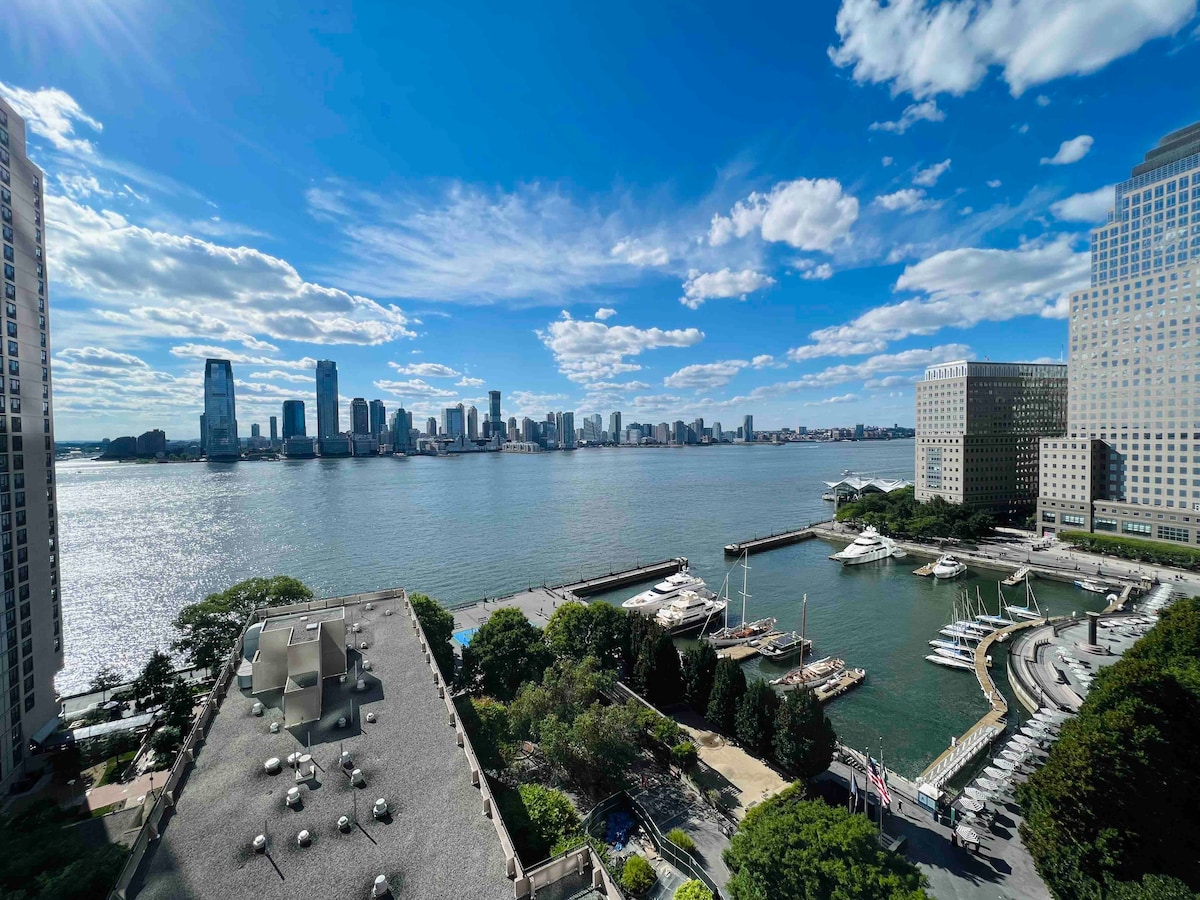 A panoramic view reveals the waterfront, with sailboats moored at the marina and the skyline of the financial district in the distance. The scene is brightened by scattered clouds and sunlight reflecting on the water.
