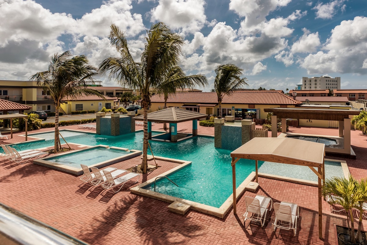 A sunny outdoor pool area features a large swimming pool with a shaded gazebo at its center. Surrounding the pool, lounge chairs are arranged for relaxation. Palm trees sway gently, and the colorful buildings of the condominium are visible in the background against a partly cloudy sky.