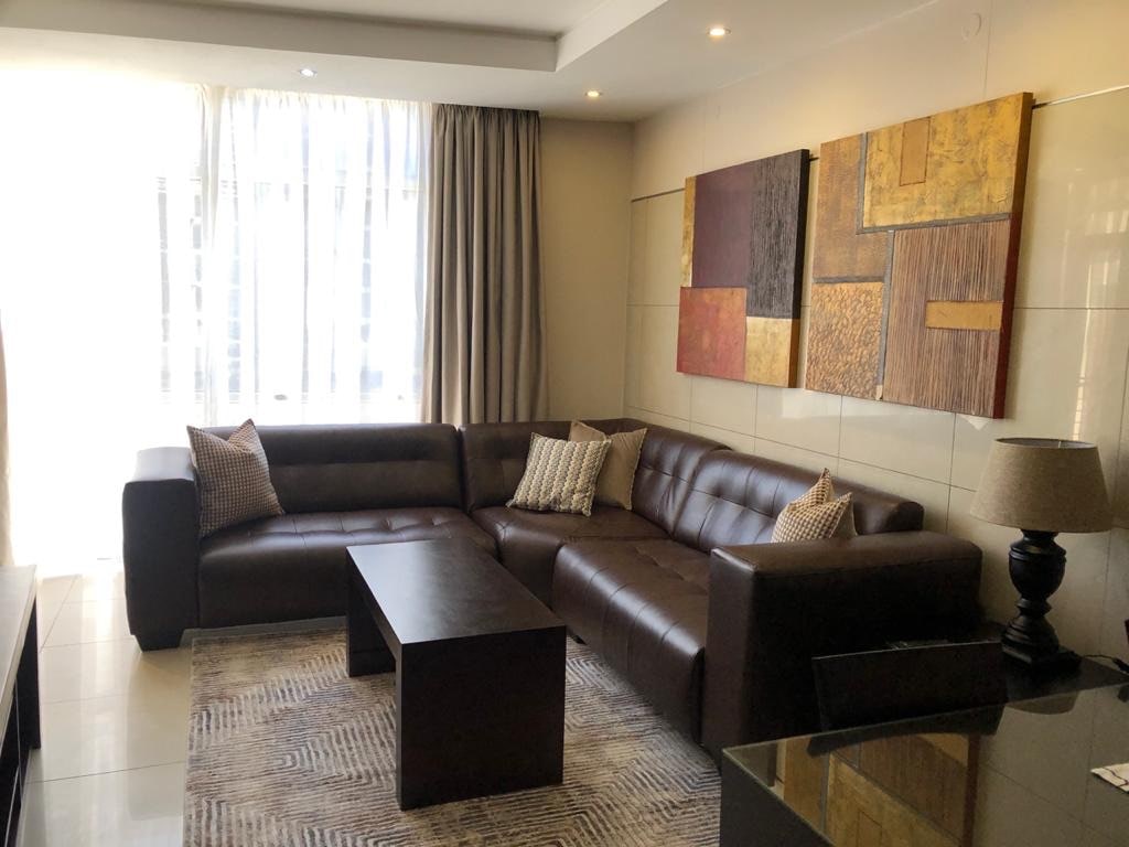 A modern living area features a brown leather sectional sofa adorned with decorative cushions. A dark wooden coffee table is positioned in front. Natural light filters through sheer curtains, illuminating a wall adorned with abstract artwork in warm tones.