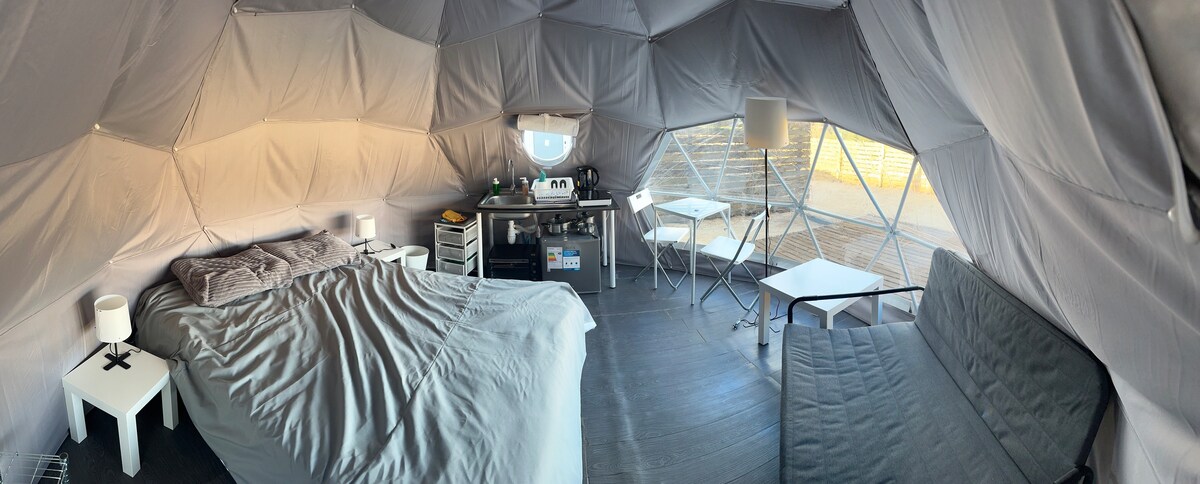 An inviting geodesic dome interior features a spacious bed covered with a soft gray blanket. A small kitchenette area is positioned nearby, alongside a dining table and folding chairs. Natural light streams in through large windows, illuminating the cozy seating area with a futon.