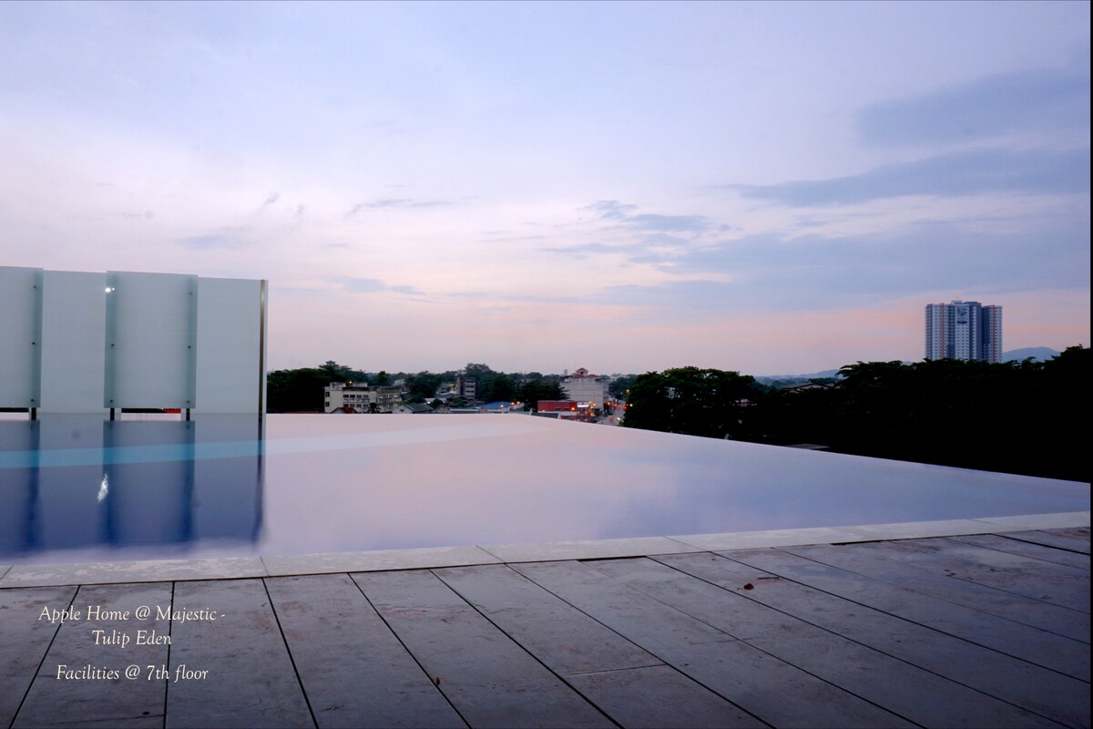 An infinity pool extends towards the horizon, reflecting the cloudy sky above. The surrounding deck is lined with smooth tiles, providing an elegant space for relaxation. A city skyline is visible in the distance, creating a serene backdrop.