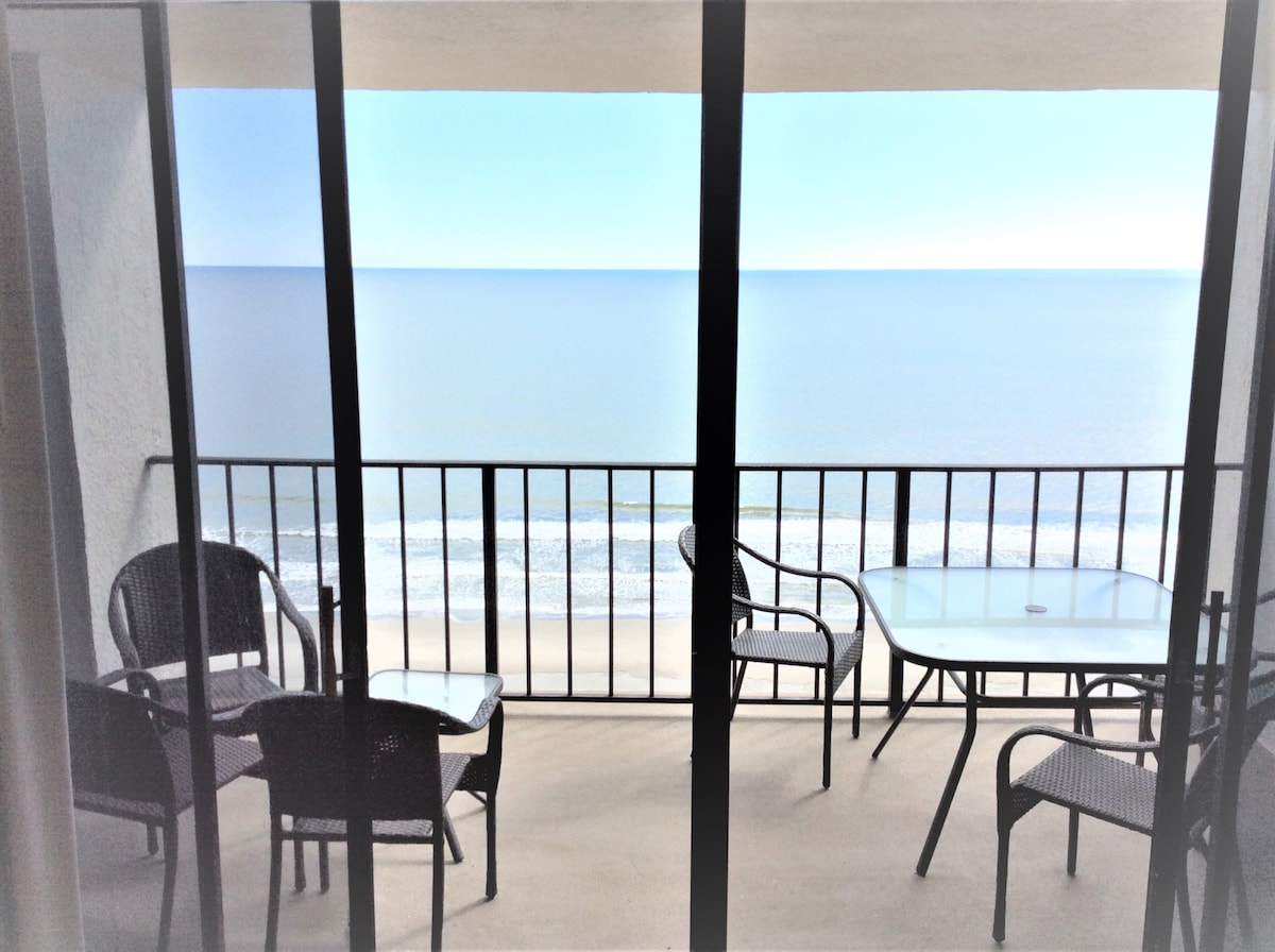 A spacious balcony offers ocean views extending to the horizon. Outdoor furniture includes a glass-top table surrounded by four chairs, providing an area for relaxation or dining. The scene is framed by glass sliding doors, highlighting the connection to indoor living space.