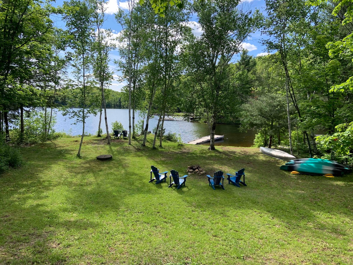 A spacious lawn area overlooks Raven Lake, surrounded by tall trees providing shade. Bright blue Adirondack chairs are arranged around a fire pit. A canoe and kayaks can be seen at the water's edge, with a gentle sloping path leading to the dock.