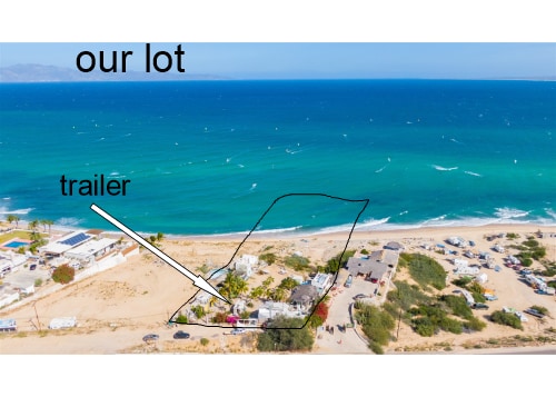An aerial view showcases the property layout with the trailer indicated. The sandy beach stretches alongside the vibrant blue ocean, contrasting with the greenery around the trailer. Nearby, other structures and vehicles can be seen, highlighting the spacious beachfront area.