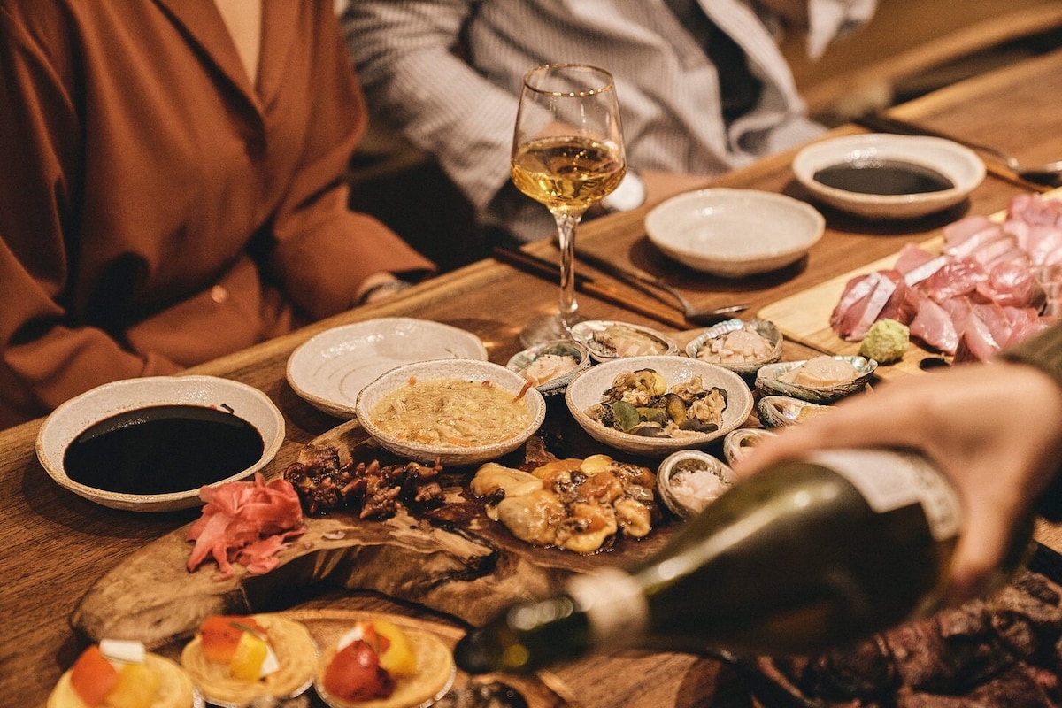 A beautifully arranged table displays an array of dishes, including sushi, seafood, and garnishes. Several plates and bowls showcase colorful ingredients, while a wine glass filled with golden liquid catches the light. The setting emphasizes a communal dining experience.