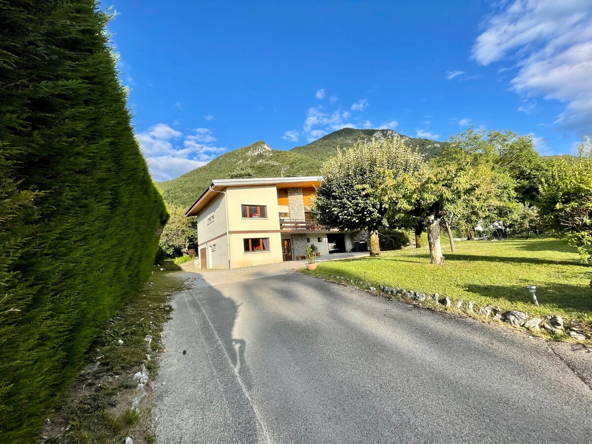 The family home is presented with a two-story structure situated on a quiet road. Lush greenery surrounds the property, and mountains rise in the background under a partly cloudy sky. A spacious lawn and fruit trees are visible in the foreground.