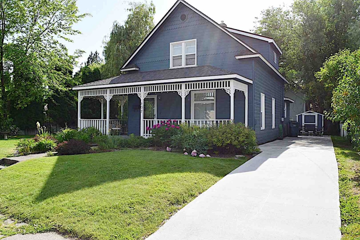 A charming blue house features a wide front porch with white trim, surrounded by lush greenery and colorful flower beds. A spacious driveway leads to the house, providing easy access to the entrance. The well-maintained yard enhances the inviting appearance of the property.