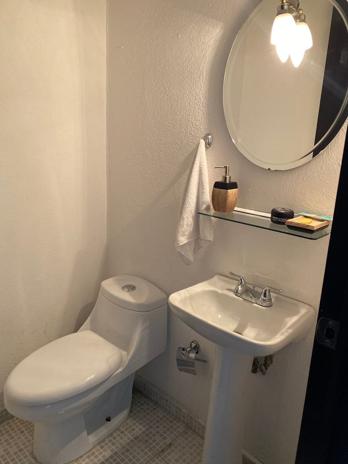 A clean and functional half bathroom is shown, featuring a toilet and a pedestal sink. A round mirror is mounted above the sink, accompanied by a small glass shelf holding toiletries. Soft lighting is provided by a ceiling fixture.