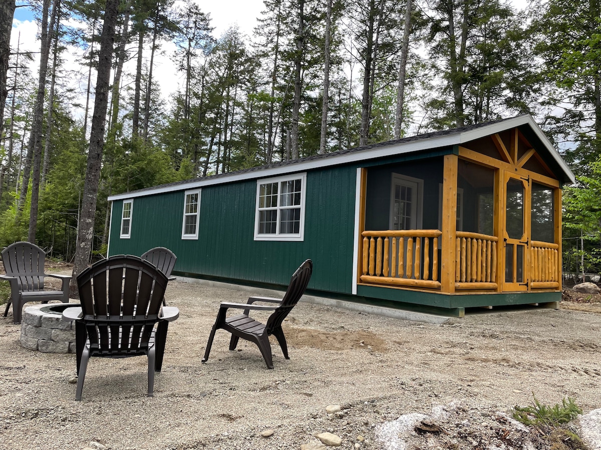 A green cabin with a screened porch is surrounded by trees, creating a private retreat. In the foreground, four black Adirondack chairs are arranged around a circular fire pit made of stone, inviting outdoor gatherings. The area is gravel, ensuring easy access.