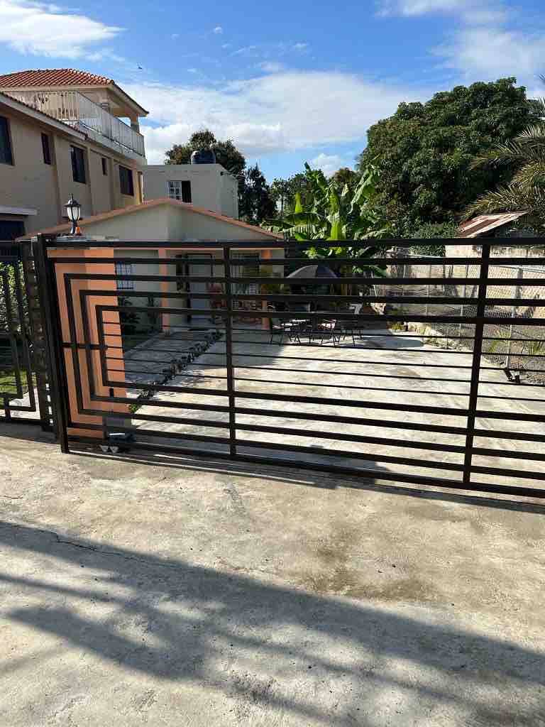 A metal gate marks the entrance to the property, revealing a spacious, paved area beyond. Shaded by surrounding trees, the grounds include a pathway leading towards the home, with a glimpse of its exterior visible in the background.