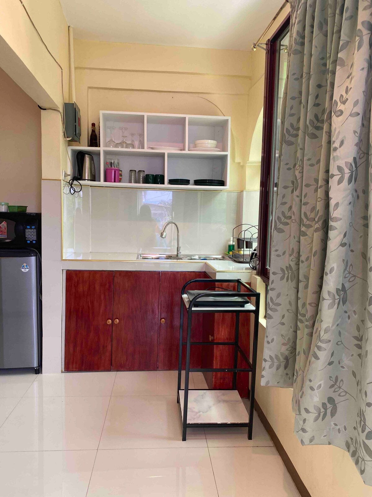 A modern kitchenette is presented with wooden cabinetry and a stainless steel sink. A small black cart offers additional storage, while a refrigerator is positioned nearby. Light-colored tiles enhance the clean and organized environment, complemented by patterned curtains framing a window.