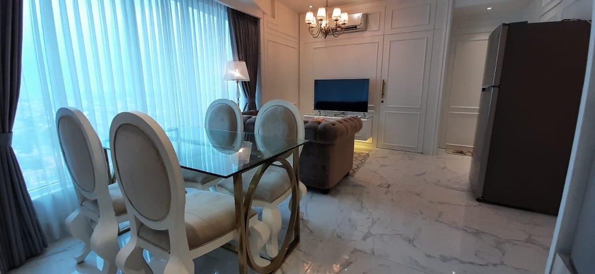 A dining area is furnished with a glass table surrounded by four elegant white chairs. The adjacent living space features a comfortable sofa and a television, with large windows allowing natural light to fill the room. The marble flooring enhances the modern aesthetic.