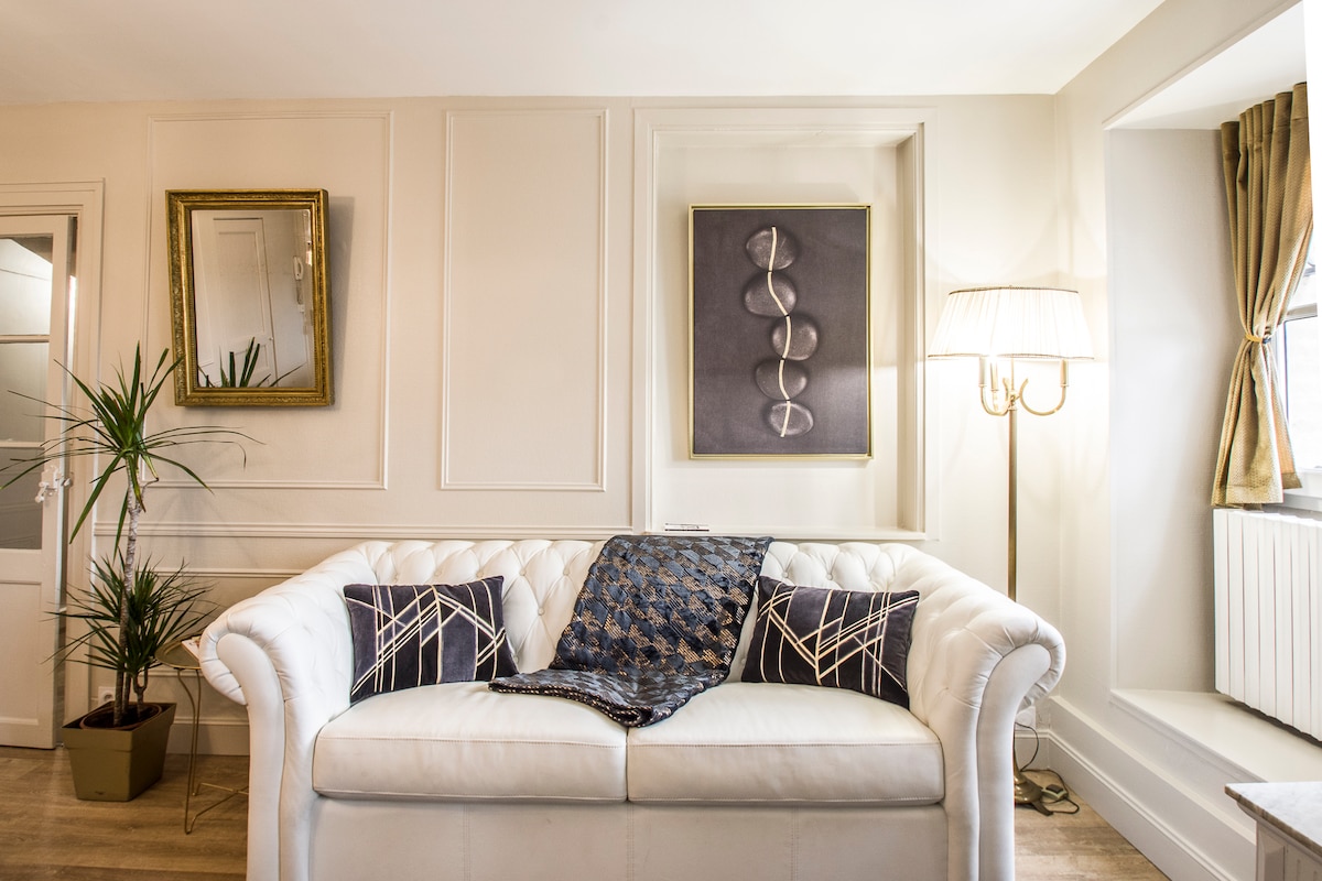 A white tufted sofa is positioned against a textured wall, accompanied by two decorative pillows. Above the sofa, a framed artwork is displayed, while a tall lamp illuminates the corner. A decorative plant adds a touch of greenery to the space.
