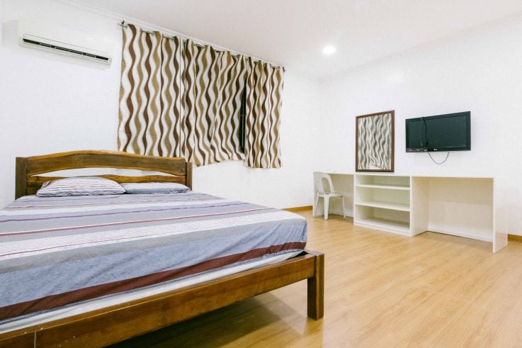A spacious bedroom features a wooden bed with striped bedding, complemented by a wall-mounted TV and a simple desk area. Natural light enters through the window, with patterned curtains adding a touch of style. The wooden floor creates a warm and inviting atmosphere.
