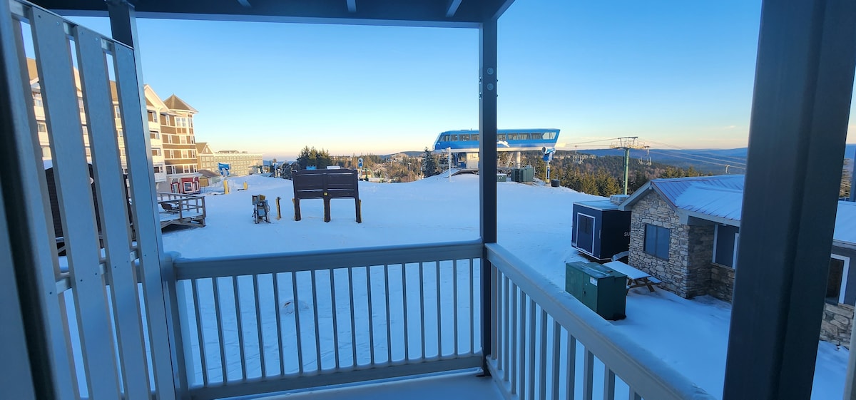 A balcony view provides a wide perspective of the snow-covered slopes and the Ballhooter chairlift in the distance. Buildings in the foreground highlight the surrounding area, while the clear sky casts soft light over the scene.