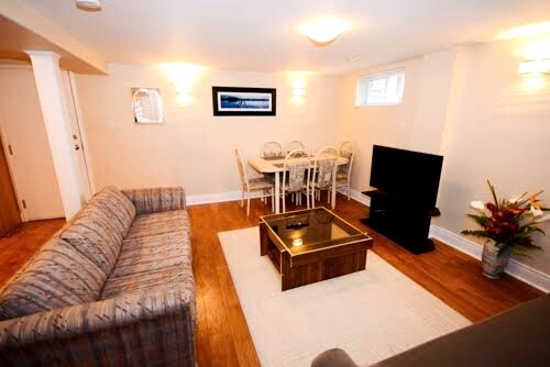 A well-lit living and dining area is featured with a comfortable sofa, a wooden coffee table, and a flat-screen television. A dining table with chairs is positioned adjacent to the living space. Soft lighting complements the warm wood flooring, enhancing the inviting ambiance.