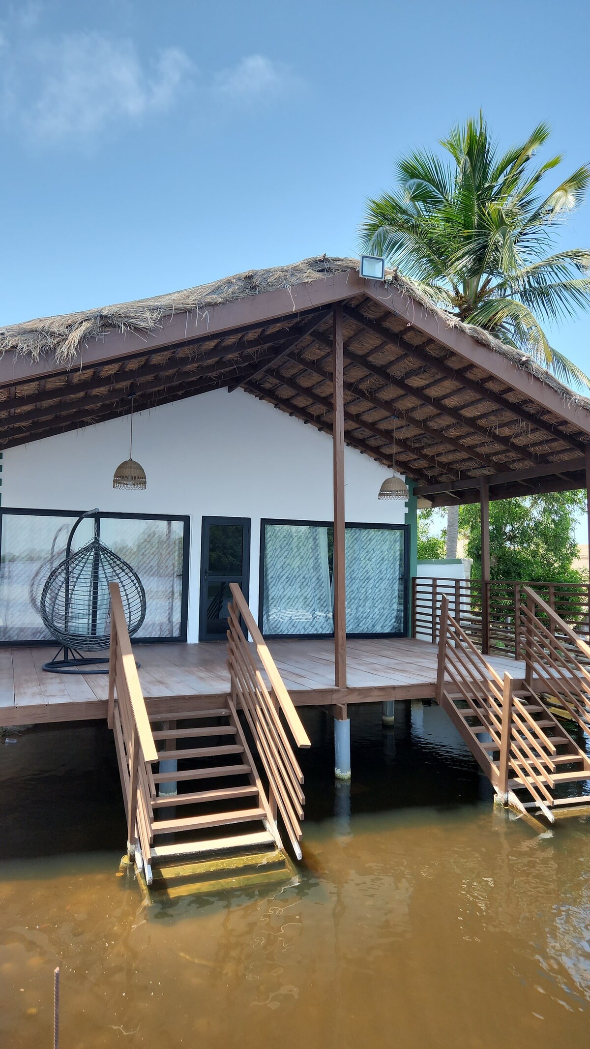 A modern cabin is positioned over the water, featuring a thatched roof and large glass doors. A stylish hanging chair is visible on the spacious deck, which is surrounded by palm trees. Stairs lead down to the serene water below.