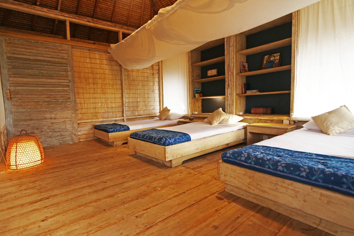 The sleeping area features three single beds with light bedding and mosquito nets, positioned on wooden platforms. Shelves with books are built into the wall, and warm lighting from a woven lantern adds a soft glow to the wooden floor and whitewashed walls.