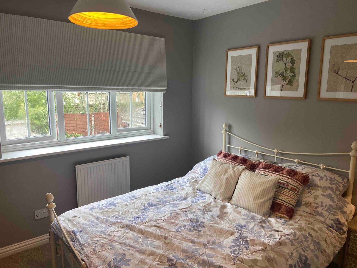 A cozy room features a double bed adorned with a floral-patterned duvet and an assortment of pillows. Light enters through a large window, accented by a shaded lamp overhead. Three botanical prints decorate the grey walls, enhancing the tranquil ambiance.
