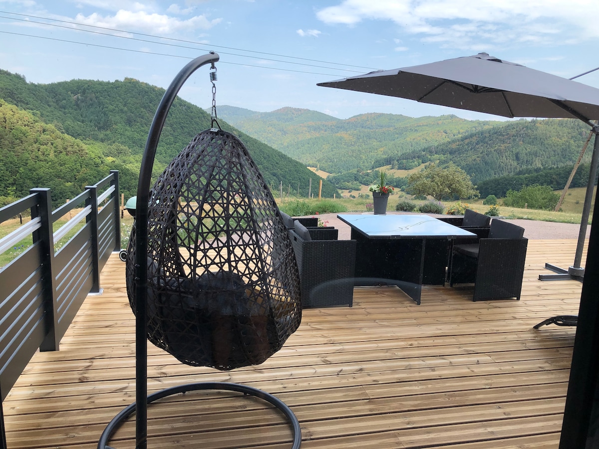 An outdoor terrace with a wooden deck features a hanging wicker chair and a dining area shaded by an umbrella. The sprawling view of lush green hills and valleys is prominent, enhancing the serene natural surroundings.