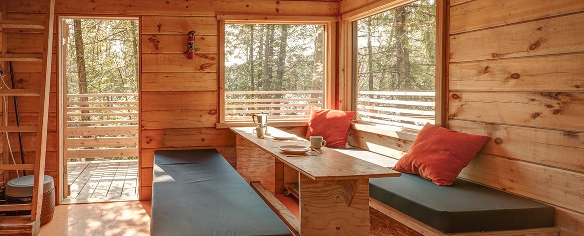 A wooden interior features large windows that allow natural light to fill the space. A table is set with dishes, and cushions are placed on benches, creating a comfortable lounging area. The warm, wooden walls contribute to a cozy atmosphere.