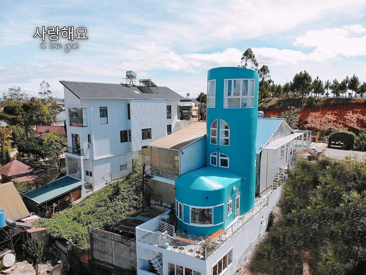 The exterior of Eudora Villa is captured from above, featuring a vibrant blue tower and surrounding structures. Lush greenery is visible, enhancing the setting. Various outdoor spaces are designed for guest enjoyment, reflecting a distinct architectural style.