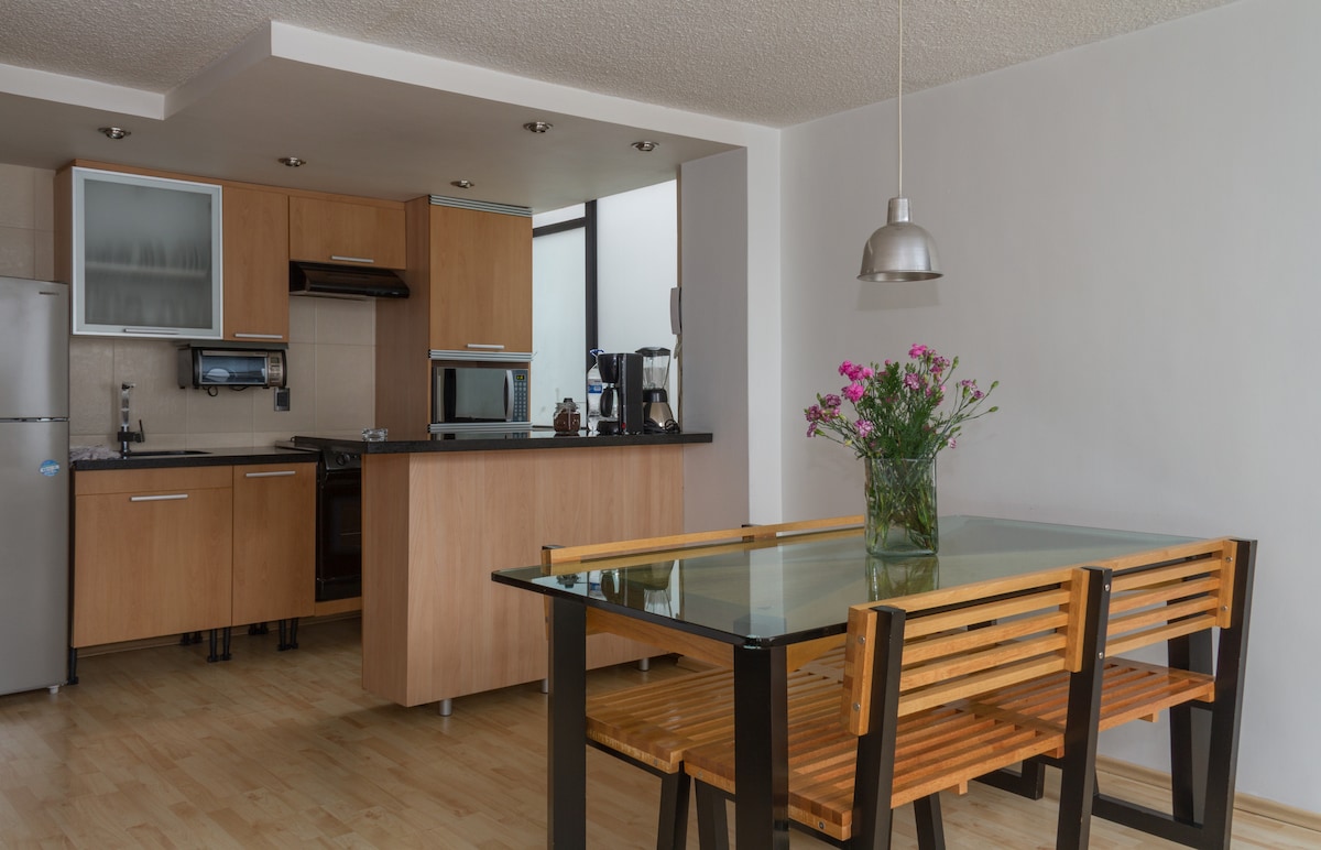 An open kitchen and dining area is showcased, featuring a glass dining table with wooden benches. The kitchen is equipped with modern appliances, including a refrigerator, microwave, and stovetop. Natural light enters from a window, highlighting the clean and minimalist design.