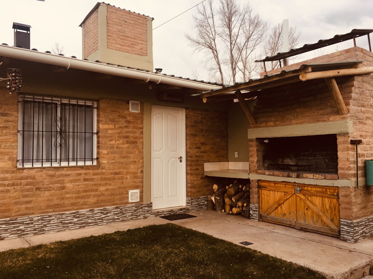 The exterior of a holiday cabin is shown, featuring rustic brick walls and a white front door. To the right, a stone barbecue area is highlighted, with a wooden door and stacked firewood beneath a covered space. A small lawn area complements the layout.