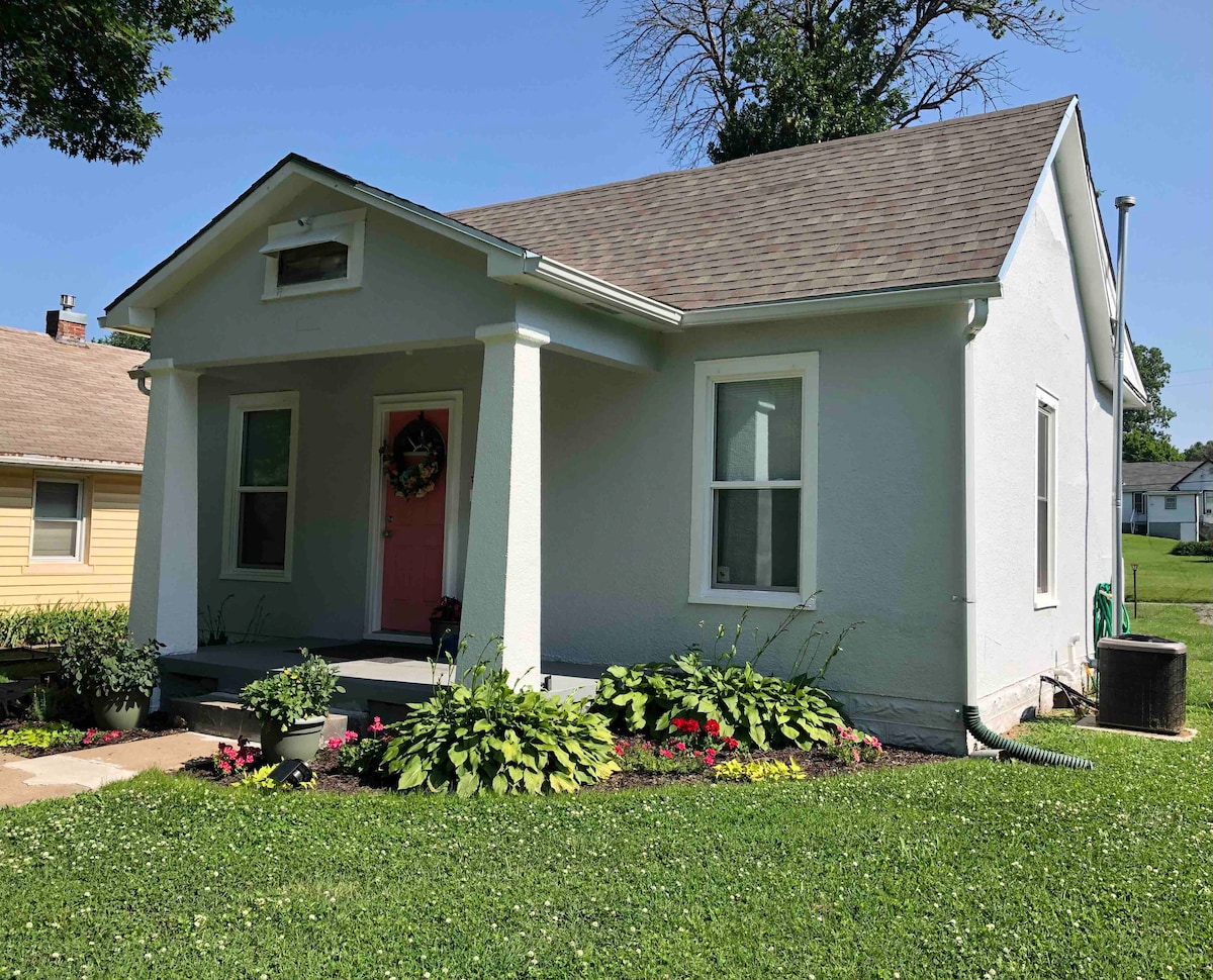 A charming bungalow is presented with a light-colored facade and a welcoming front porch. The entrance features a vibrant door, while well-maintained flower beds with colorful plants adorn the surrounding area. A lush green lawn completes the inviting exterior.
