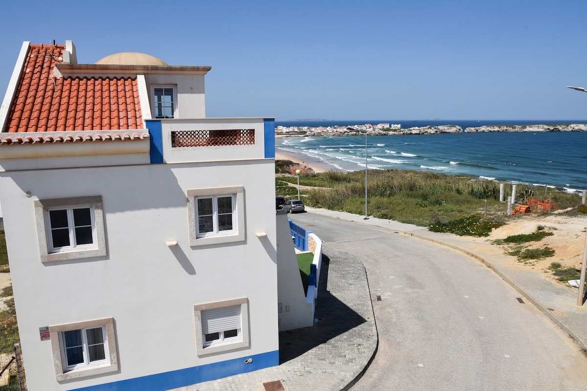 The exterior of a two-story house displays a charming coastal design, with a terracotta roof and white walls accented by blue trim. The sweeping view of the ocean and beach can be seen from the property, framed by gentle waves and a clear sky.