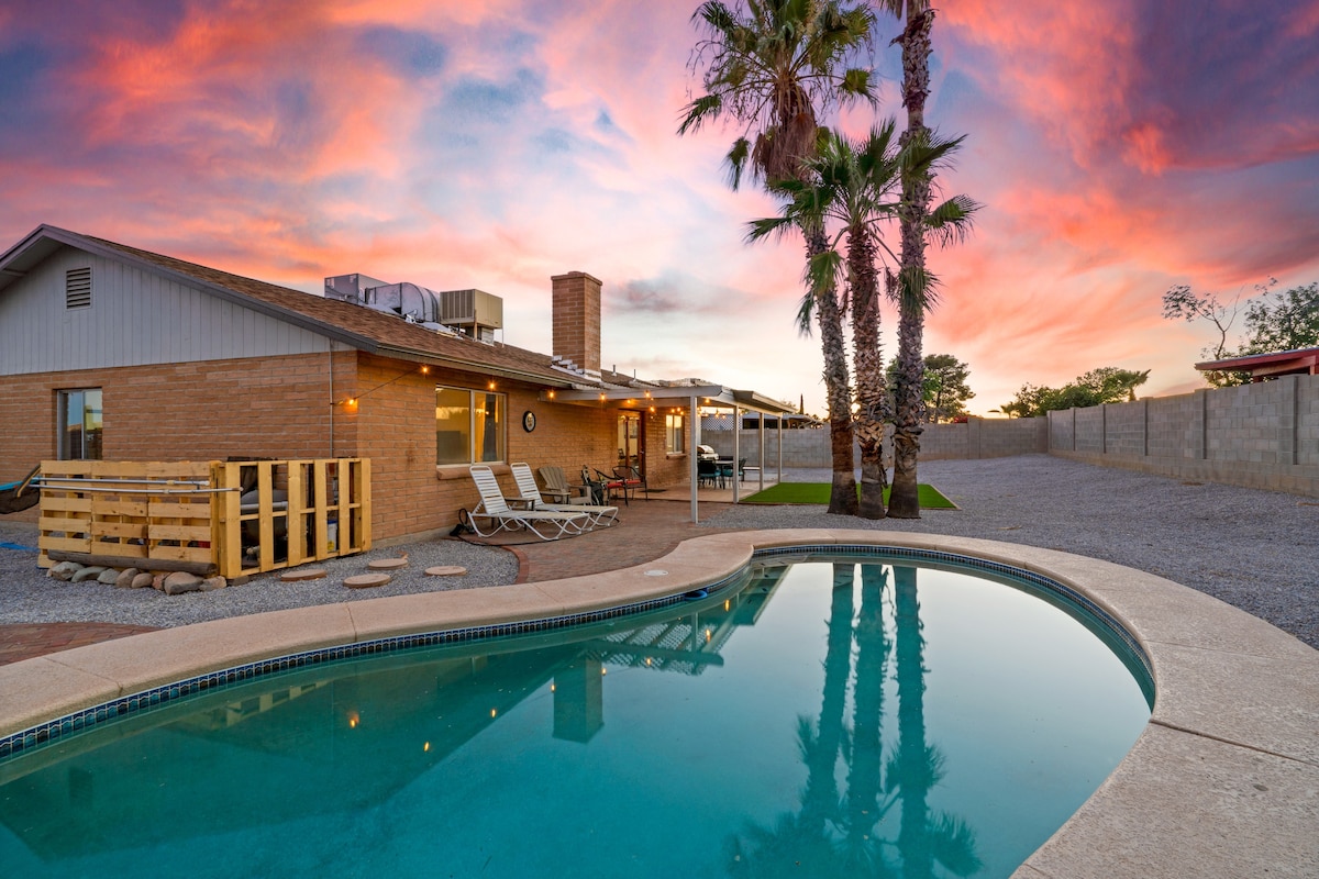 A serene backyard scene features a shimmering pool reflecting the vibrant sunset sky. Palm trees stand beside the pool, and lounge chairs are arranged nearby. A covered patio extends from the house, inviting relaxation and offering an unobstructed view of the desert landscape.
