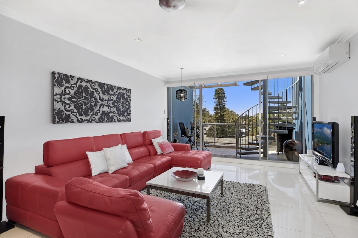 The lounge area features a spacious red leather modular sofa and a minimalist coffee table atop a textured rug. Large glass doors open to a balcony, allowing natural light and showcasing views of the outdoor space. A flat-screen TV is positioned in the corner.