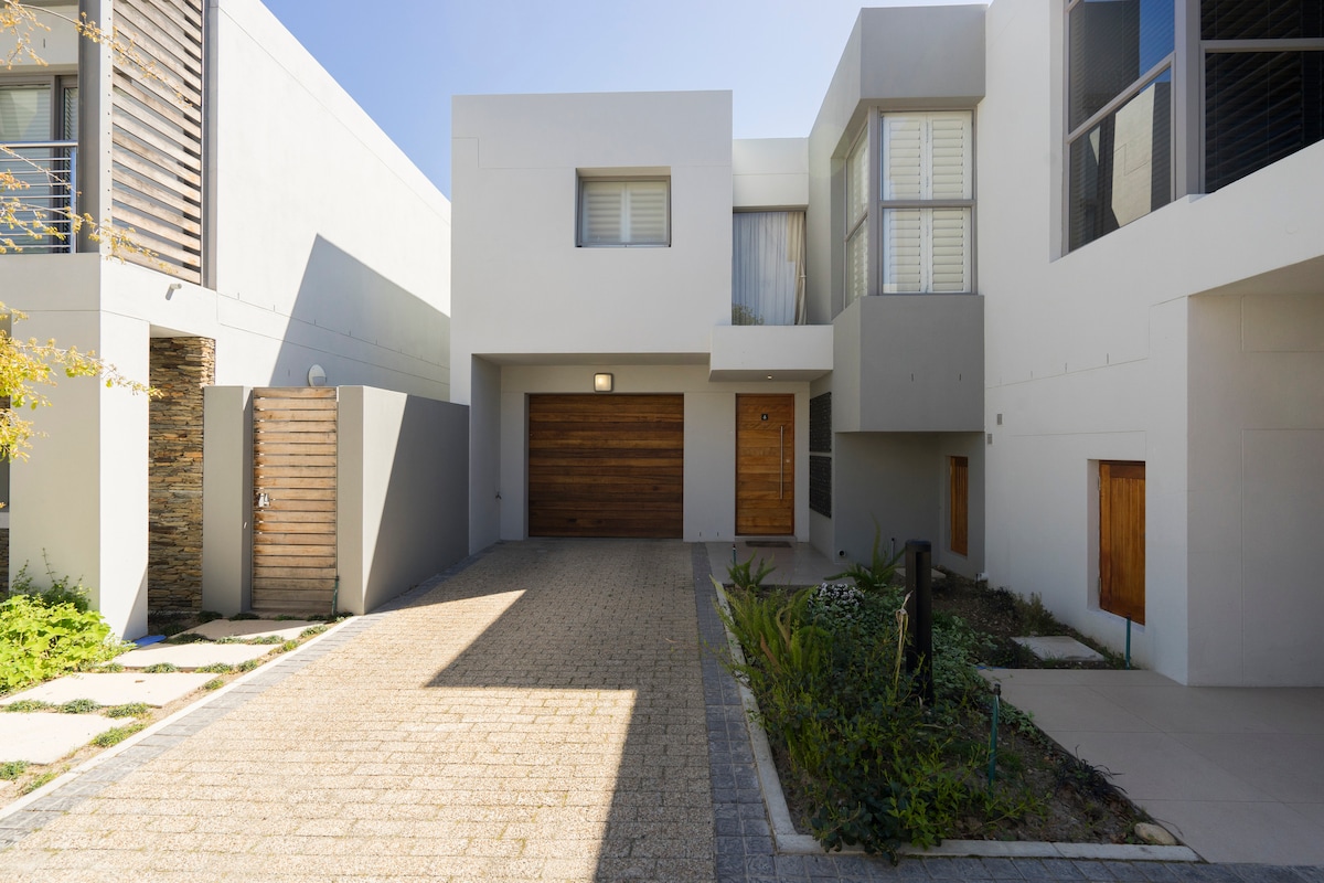 A modern, multi-story building is showcased, featuring a blend of wood and gray elements. A spacious driveway made of cobblestones leads to the entrance, framed by neatly manicured greenery and contemporary architecture.