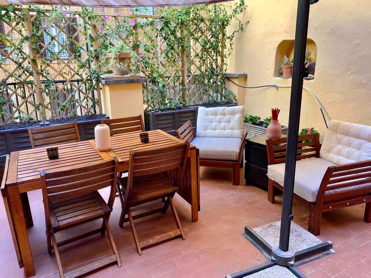 An outdoor terrace is furnished with a wooden dining table and six folding chairs. Two cushioned seating areas are arranged for relaxation. Greenery climbs the trellis, adding a touch of nature to the space, while pot plants are positioned against the walls.