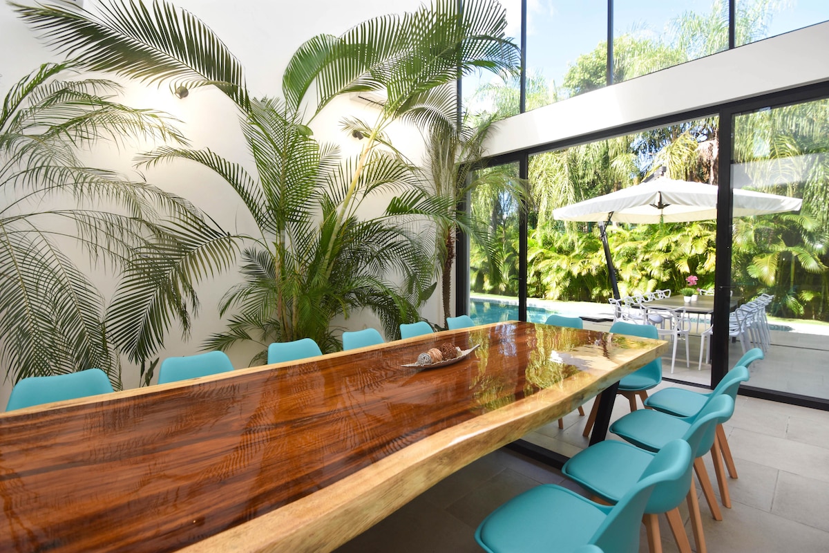 A large dining table crafted from natural wood is surrounded by mint-colored chairs. Lush greenery is featured in the background, enhanced by abundant natural light streaming in through floor-to-ceiling glass doors, creating a bright and airy ambiance.