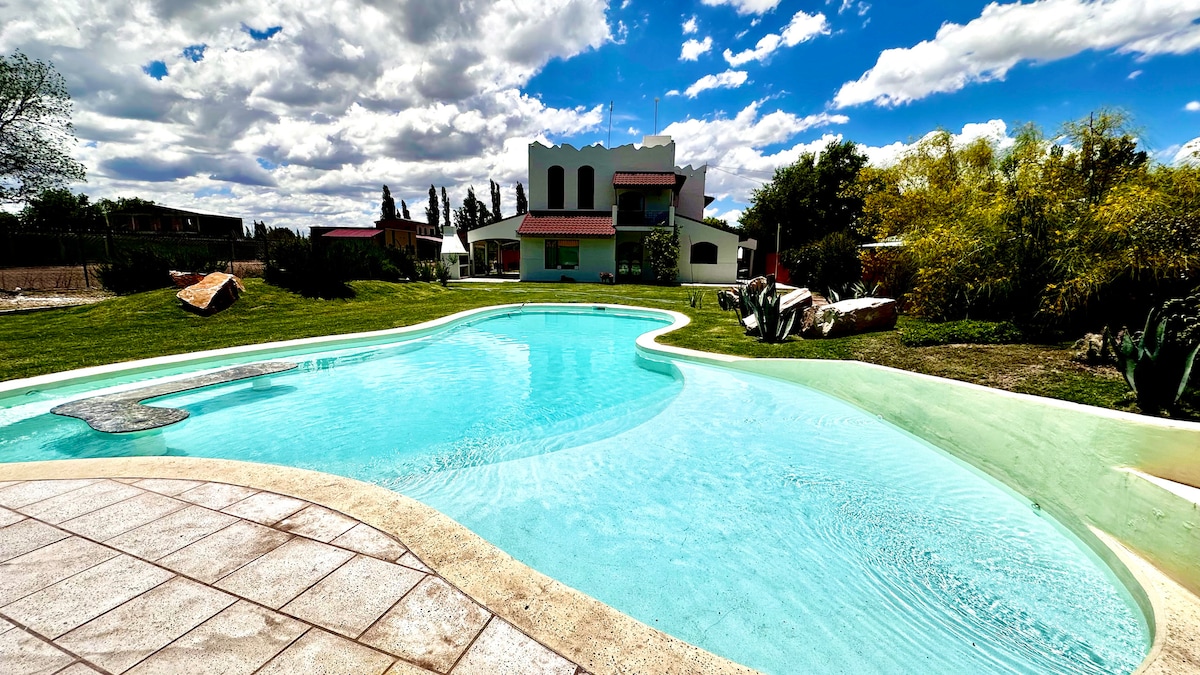 A spacious pool area is surrounded by well-maintained grass and natural landscaping. The house is visible in the background, featuring a mix of white and color accents. Bright clouds drift across a blue sky, enhancing the serene outdoor setting.