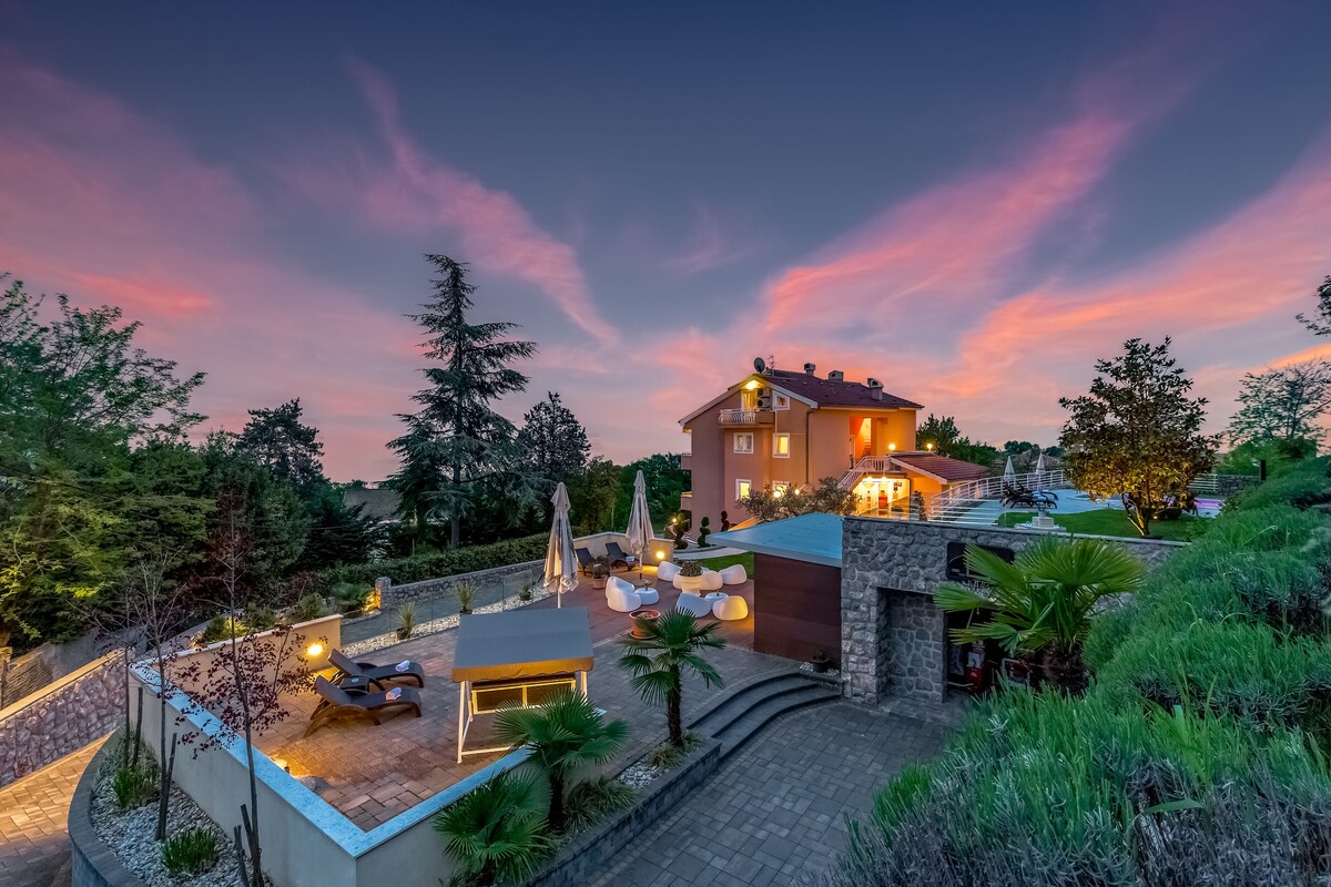 The exterior of Villa Nada is illuminated at dusk, featuring a spacious terrace with a swimming pool. A barbecue area and various seating arrangements are visible among manicured gardens. The backdrop showcases trees and a colorful sky, enhancing the tranquil ambiance of the setting.