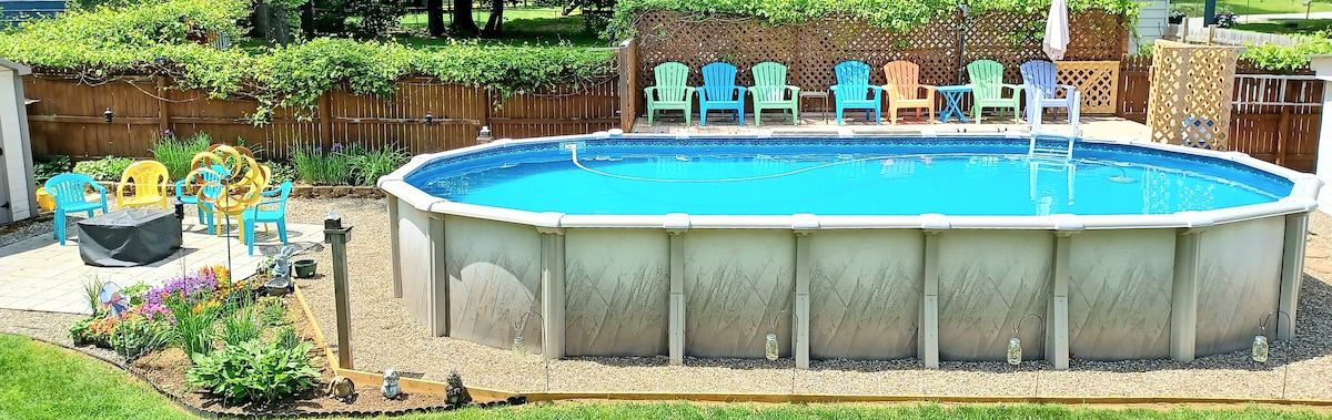 An above-ground pool is surrounded by a spacious patio area. Colorful lounge chairs are arranged nearby, creating a relaxed atmosphere. A gas fire pit sits on the patio, with decorative flowers enhancing the outdoor setting.