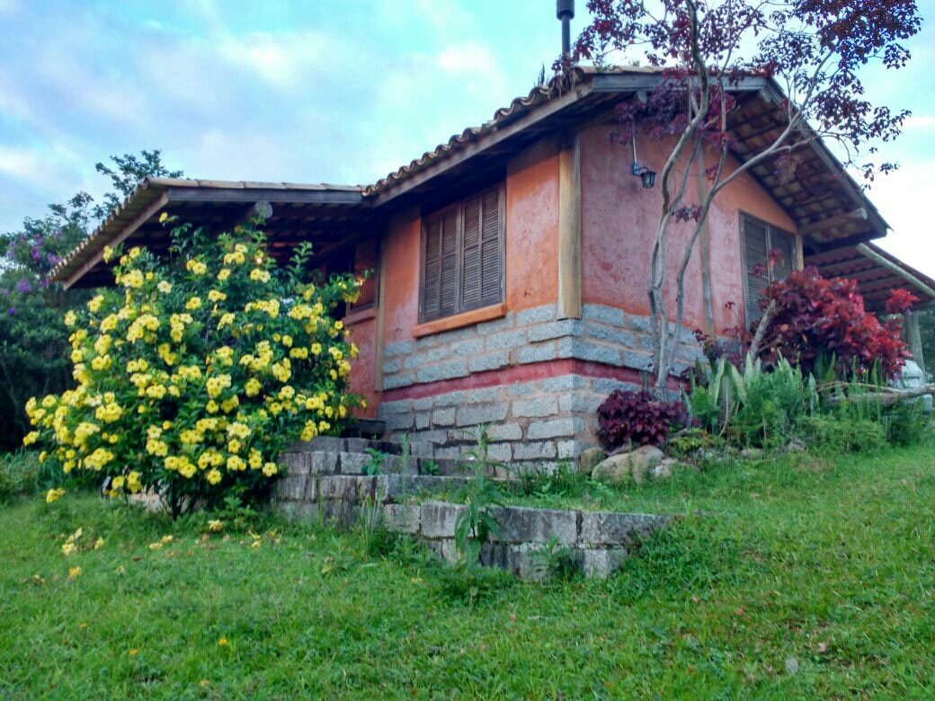 A charming cottage is nestled among lush greenery, featuring a stone foundation and wooden accents. Bright yellow flowers bloom near the entrance, while various plants adorn the landscaping. The cottage is surrounded by a tranquil rural environment, complementing the natural beauty of the area.