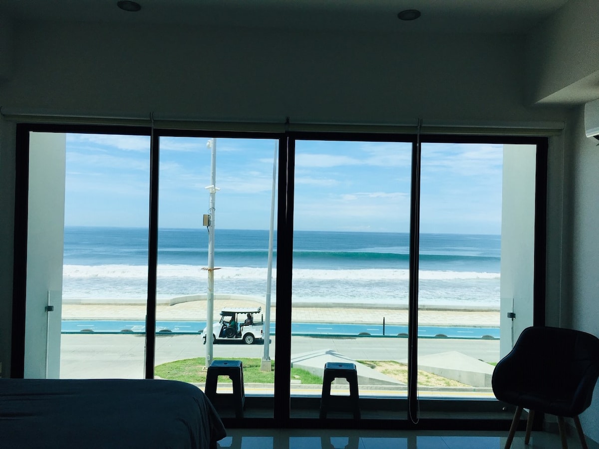 Large glass doors frame a view of the ocean, showcasing the gentle waves and beachfront beyond. A serene atmosphere is created by the natural light streaming in. The foreground features modern furniture with minimalist design elements.