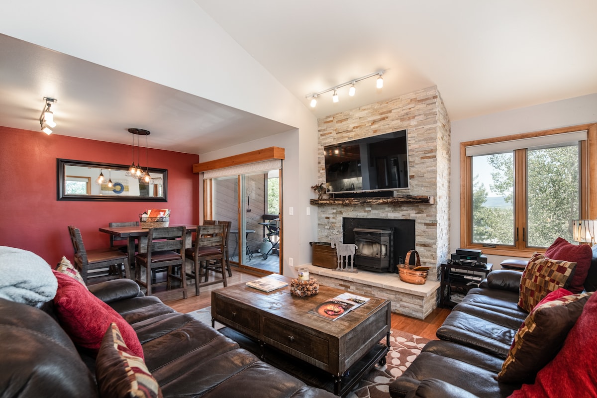 An open living area presents a blend of comfort and modernity, featuring a cozy seating arrangement with dark leather sofas, a coffee table, and a stone fireplace. Natural light enters through large windows, while a dining area with a wooden table complements the space.