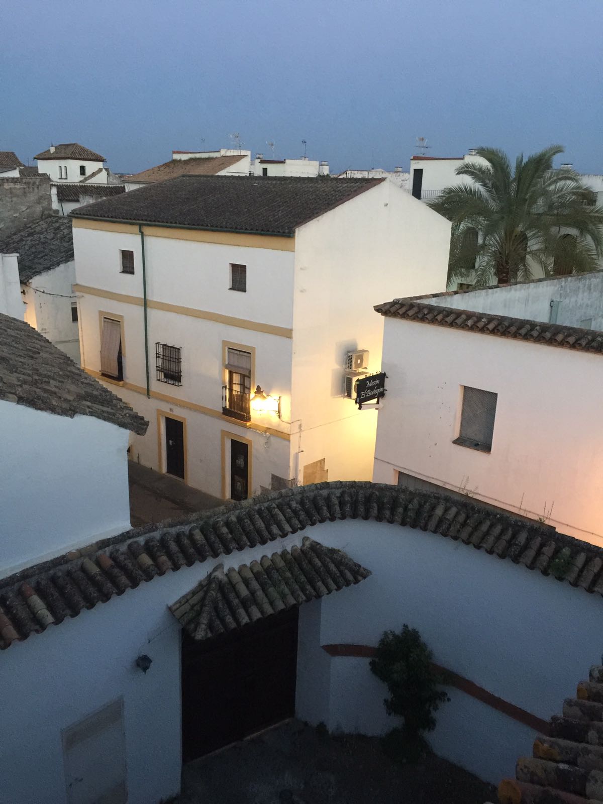 A view of traditional white buildings with tiled roofs can be seen, under a soft evening sky. Warm lights illuminate the exterior of one building, creating a cozy atmosphere amidst the peaceful surroundings of a historic neighborhood.