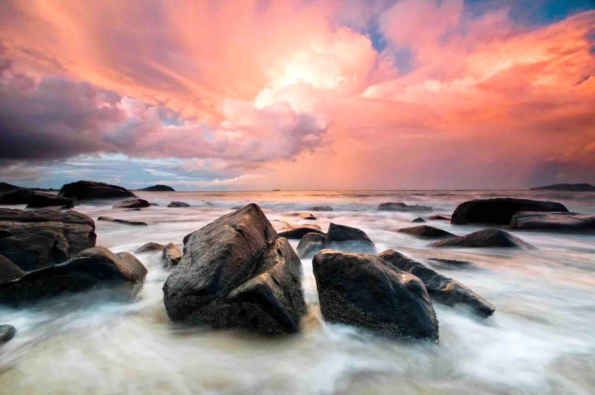 A serene seascape features smooth, dark rocks partially submerged in gentle waves. The sky is painted with shades of pink and orange, reflecting a beautiful sunset. The water appears calm as it laps against the rocks, creating a peaceful coastal atmosphere.