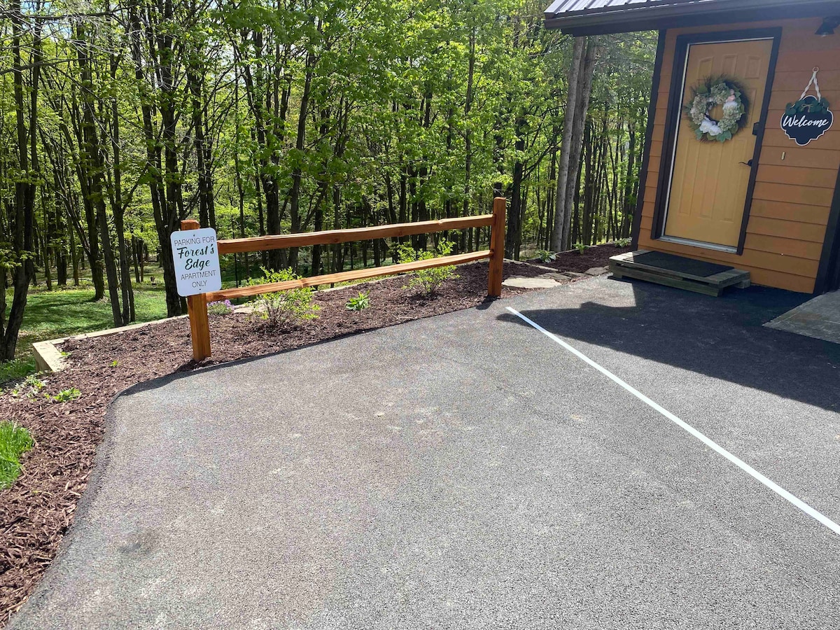 A neatly paved parking area is visible, leading to a welcoming entrance adorned with a seasonal wreath. A sign indicating "Forest's Edge" is positioned beside the parking space, and greenery from surrounding trees adds a natural backdrop to the setting.