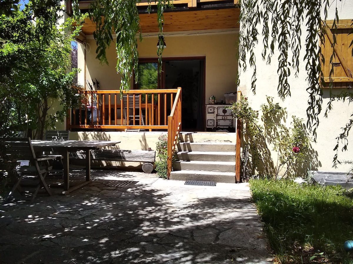 A covered terrace features wooden railings and steps leading to the entrance. A picnic table is positioned on the stone-paved area, surrounded by greenery. Flowering plants and shrubs add to the tranquil ambiance of the entryway.